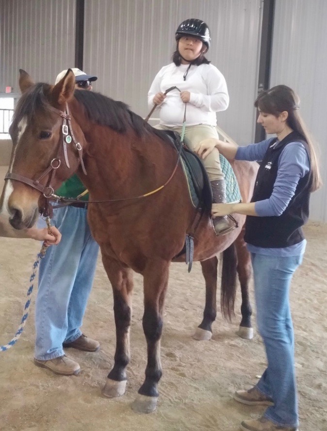 Female student riding a horse assisted by arena staff. 