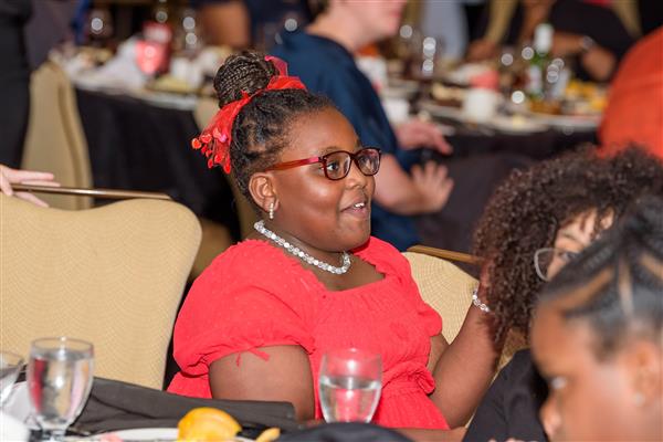 A young girl dressed in red with pearl earrings and a pearl necklace looks at the action on stage. 